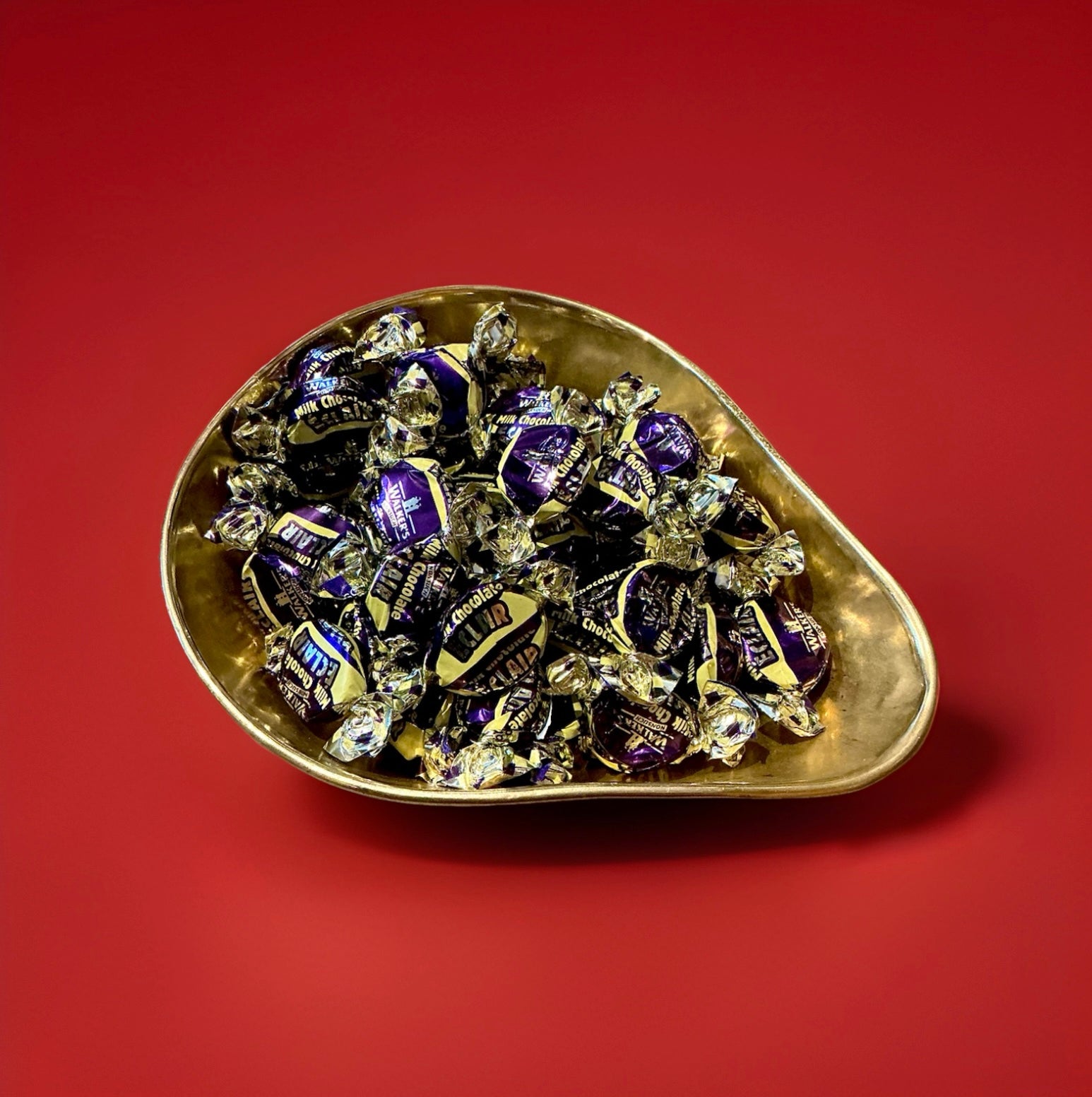 Individually wrapped purple and gold chocolate eclairs in a gold bowl on a red background