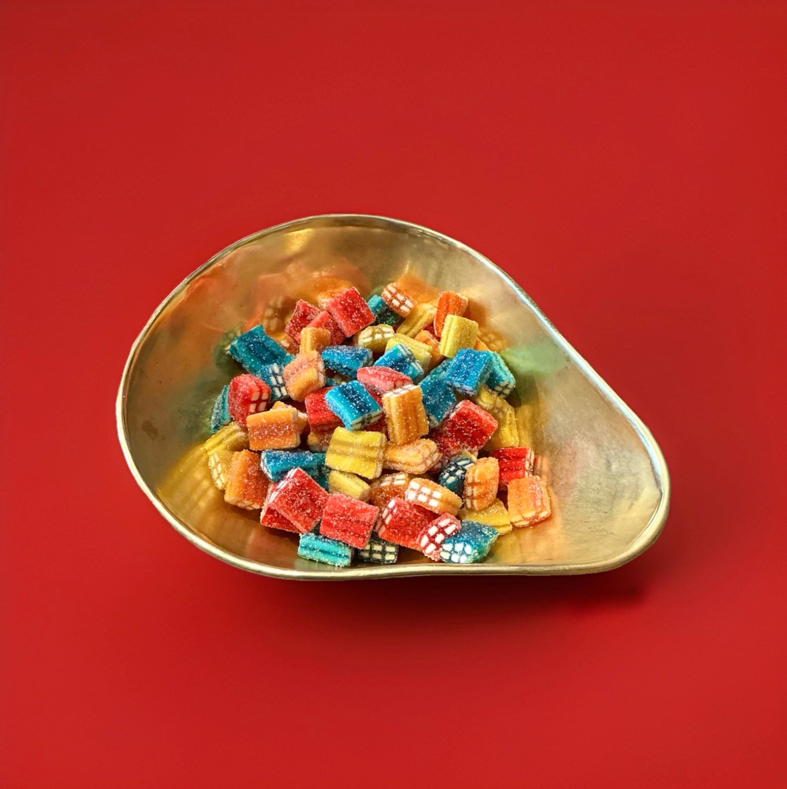 Multicoloured fruit flavoured jelly sweets in the shape of a brick with a white fondant centre and a fizzy sugar coating in a gold bowl with a red background 