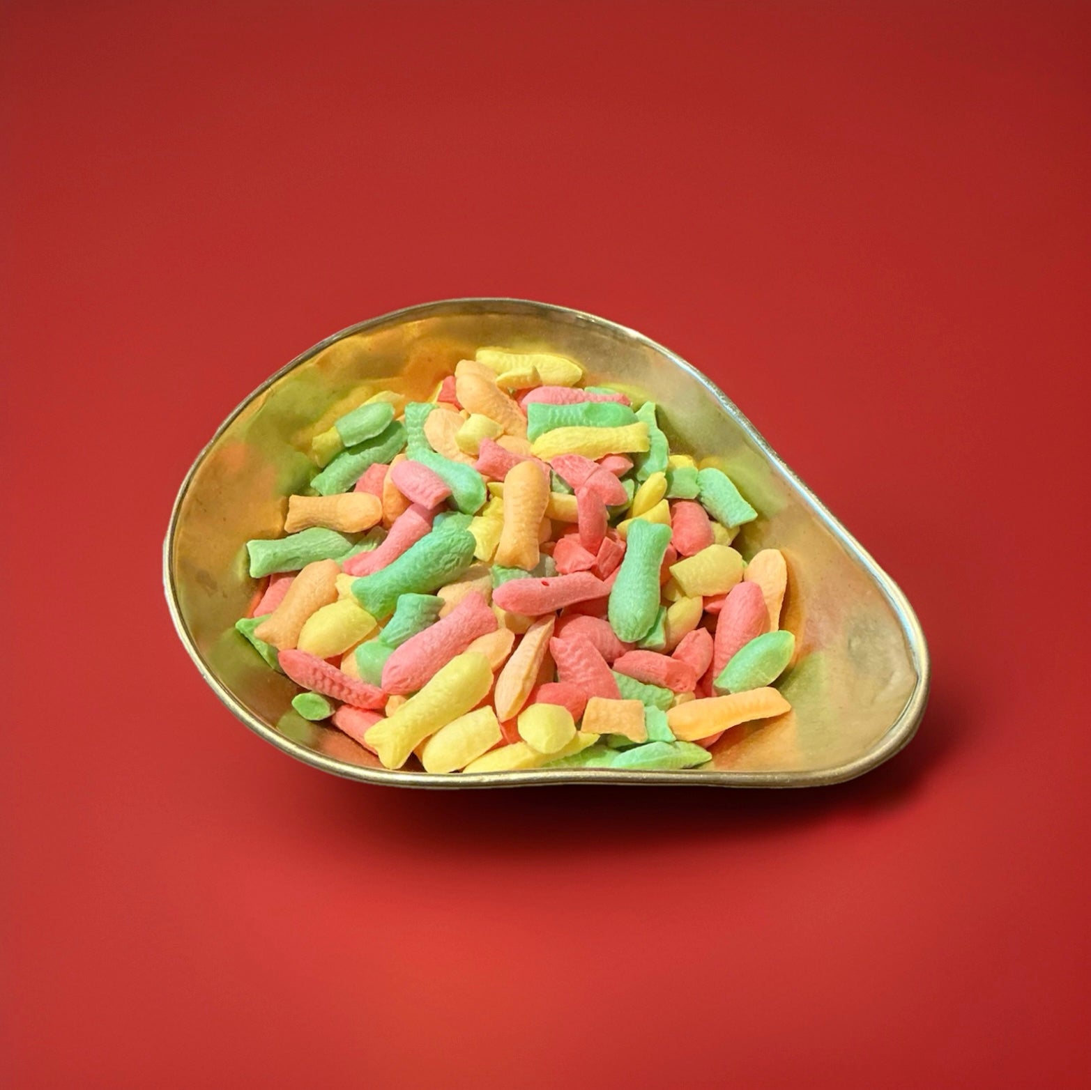 100 grams of fish shaped multicoloured hard boiled fizzy fruit sweets in a gold bowl with a red background 