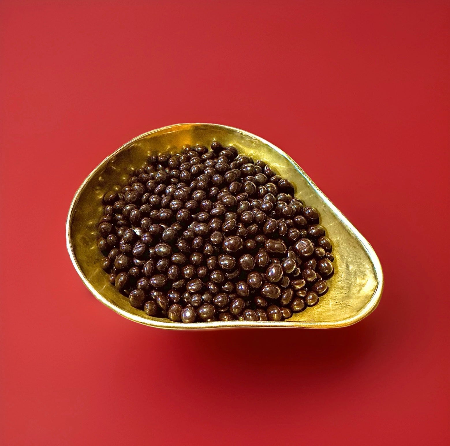 Small round bite sized pieces of Carol Anne cinder toffee coated in rich dark chocolate in a gold bowl with a red background 