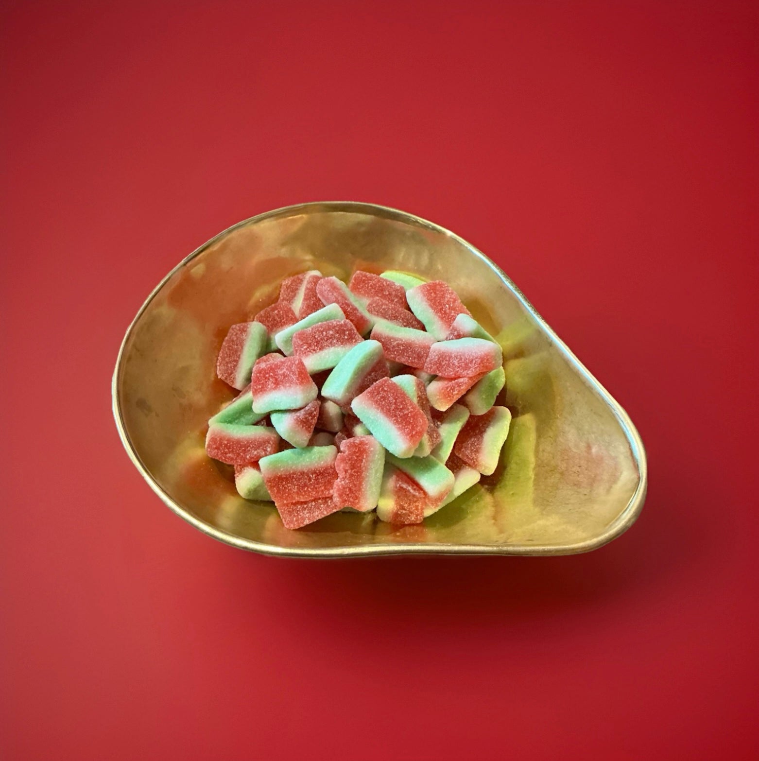 Green white and red watermelon slice shaped jelly sweets with a watermelon flavour and a fizzy sugar coating in a gold bowl with a red background 