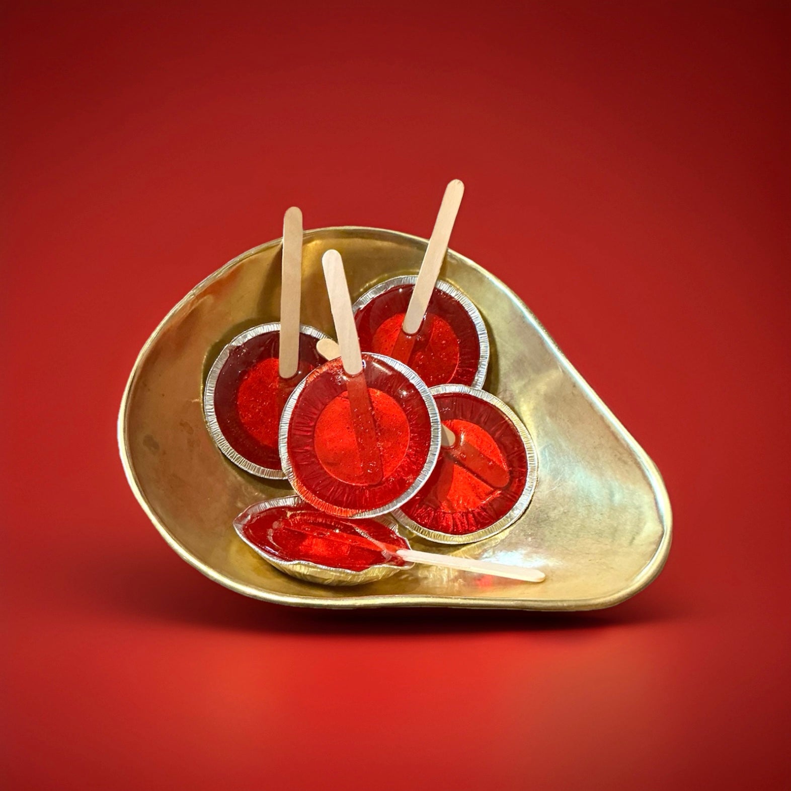 Large round hard boiled red toffee lolly in a foil cup with a wooden lolly stick in a gold bowl with a red background 