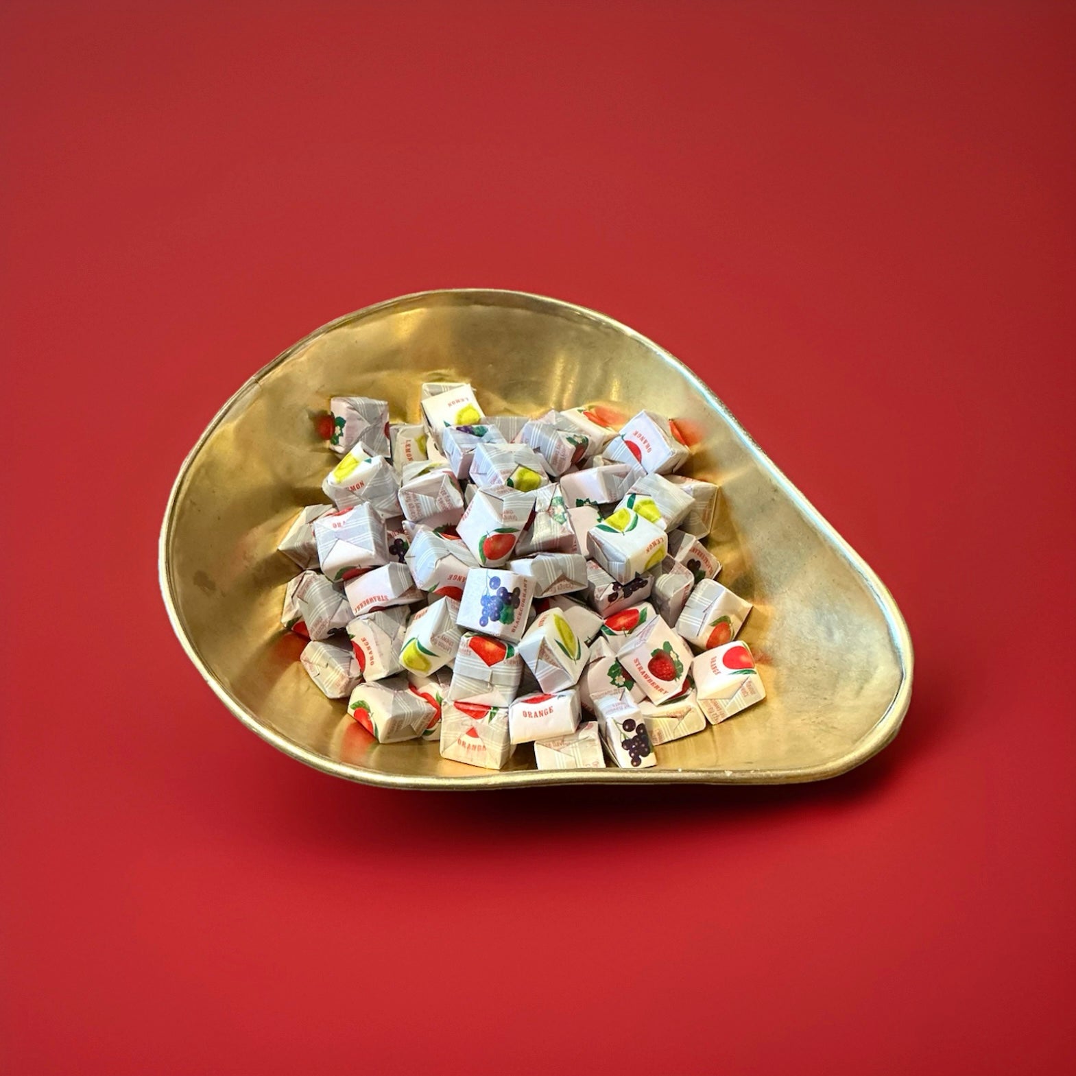 Individually wrapped fruit caramel sweets in a white wrapper with a picture of the fruit on in a gold bowl on a red background 