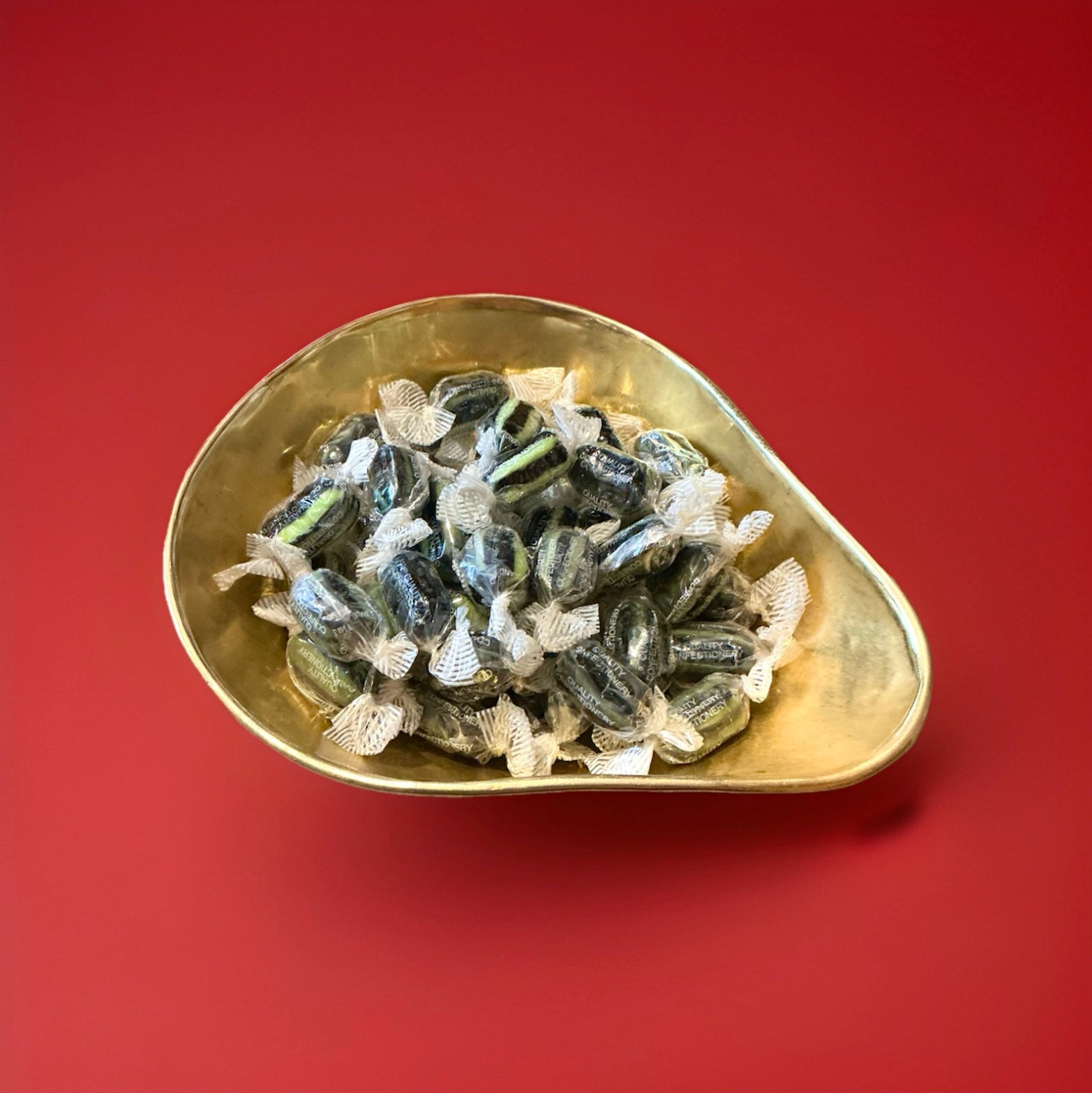 Individually wrapped liquorice and lime flavoured black and green striped sweets in a gold bowl on a red background 