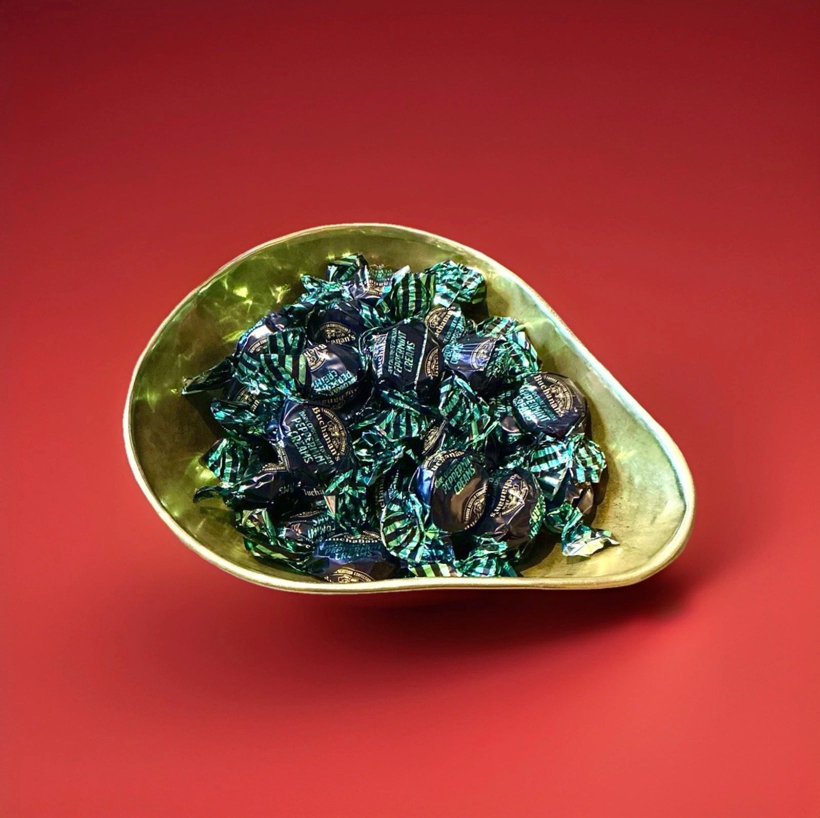 Individually wrapped peppermint flavoured fondant with a dark chocolate coating in a green wrapper in a gold bowl on a red background 