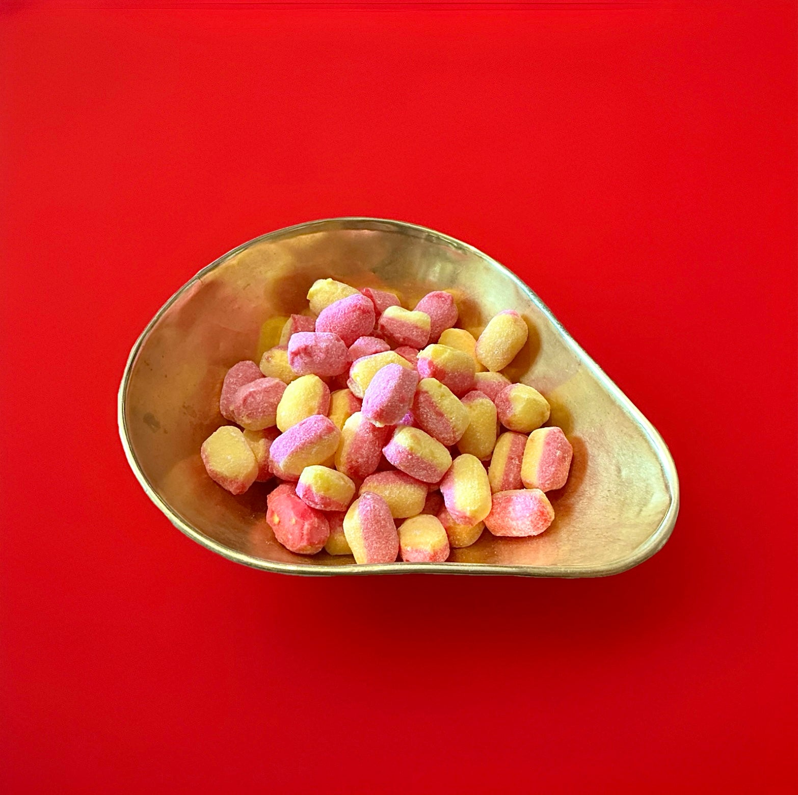 Oval half pink half yellow hard boiled rhubarb and custard sweets in a gold bowl with a red background 